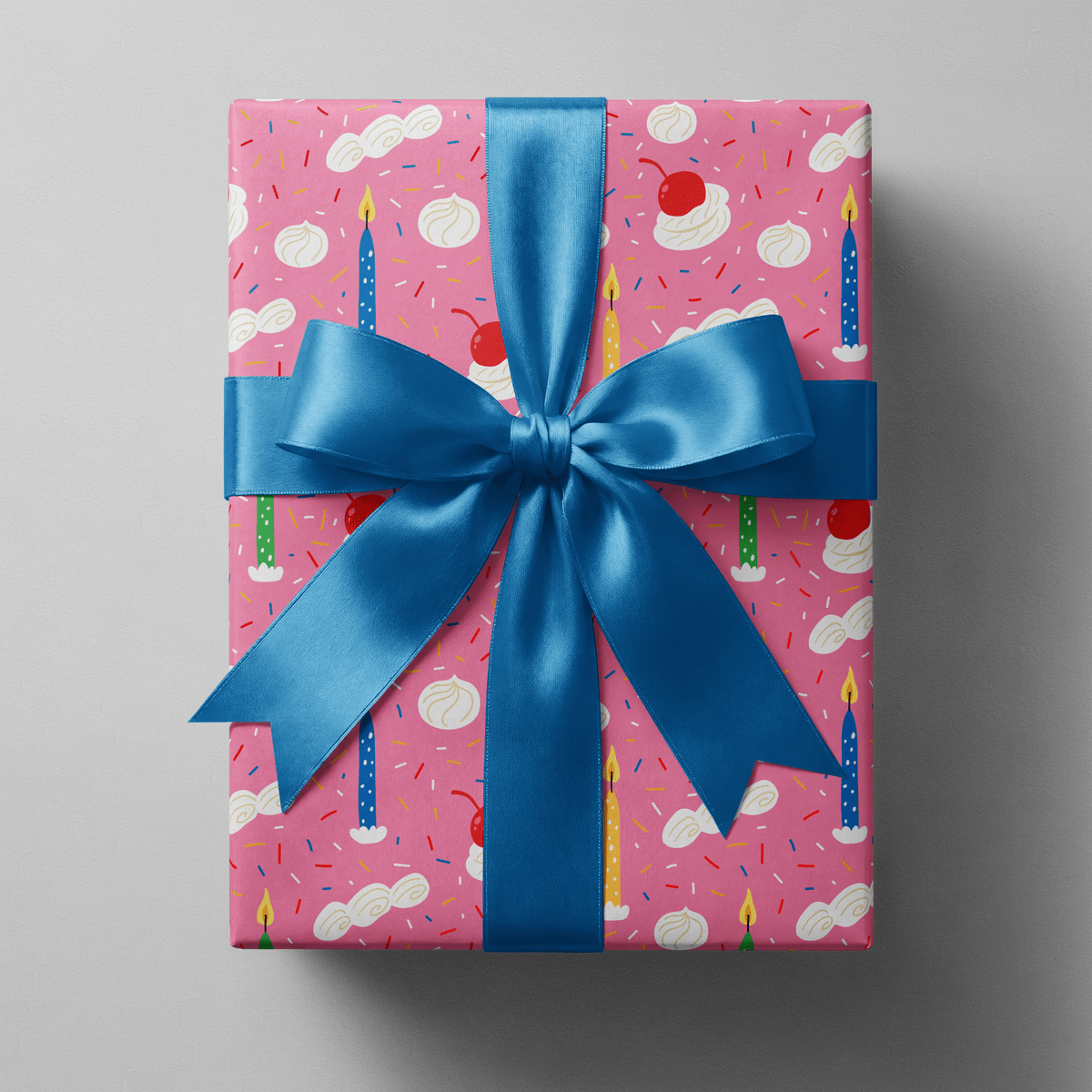 Birthday cake wrapping paper with lit candles, whipped cream swirls, and cherries on pink.