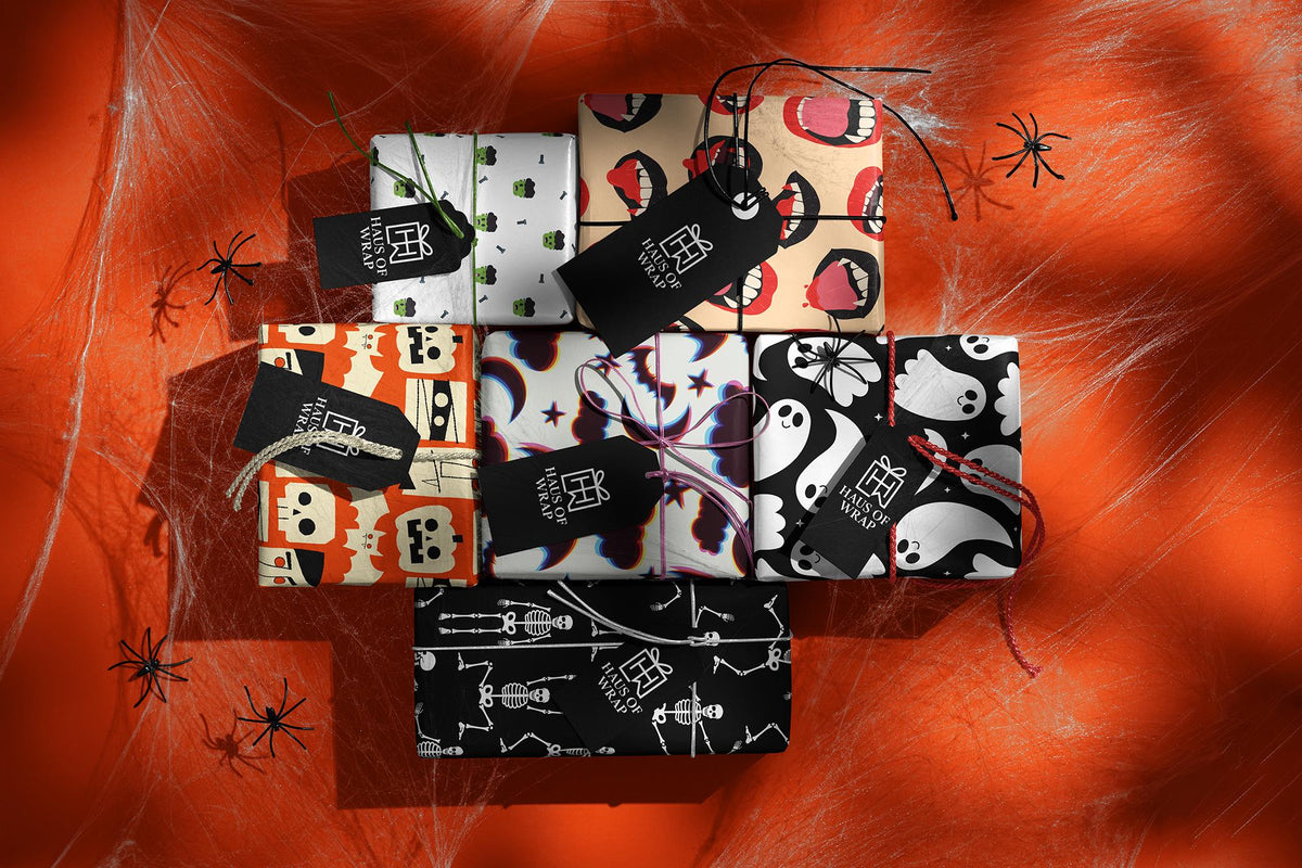 Halloween gifts wrapped in ghost, skeleton, vampire lips and pumpkin papers with Haus of Wrap tags on an orange cobweb backdrop